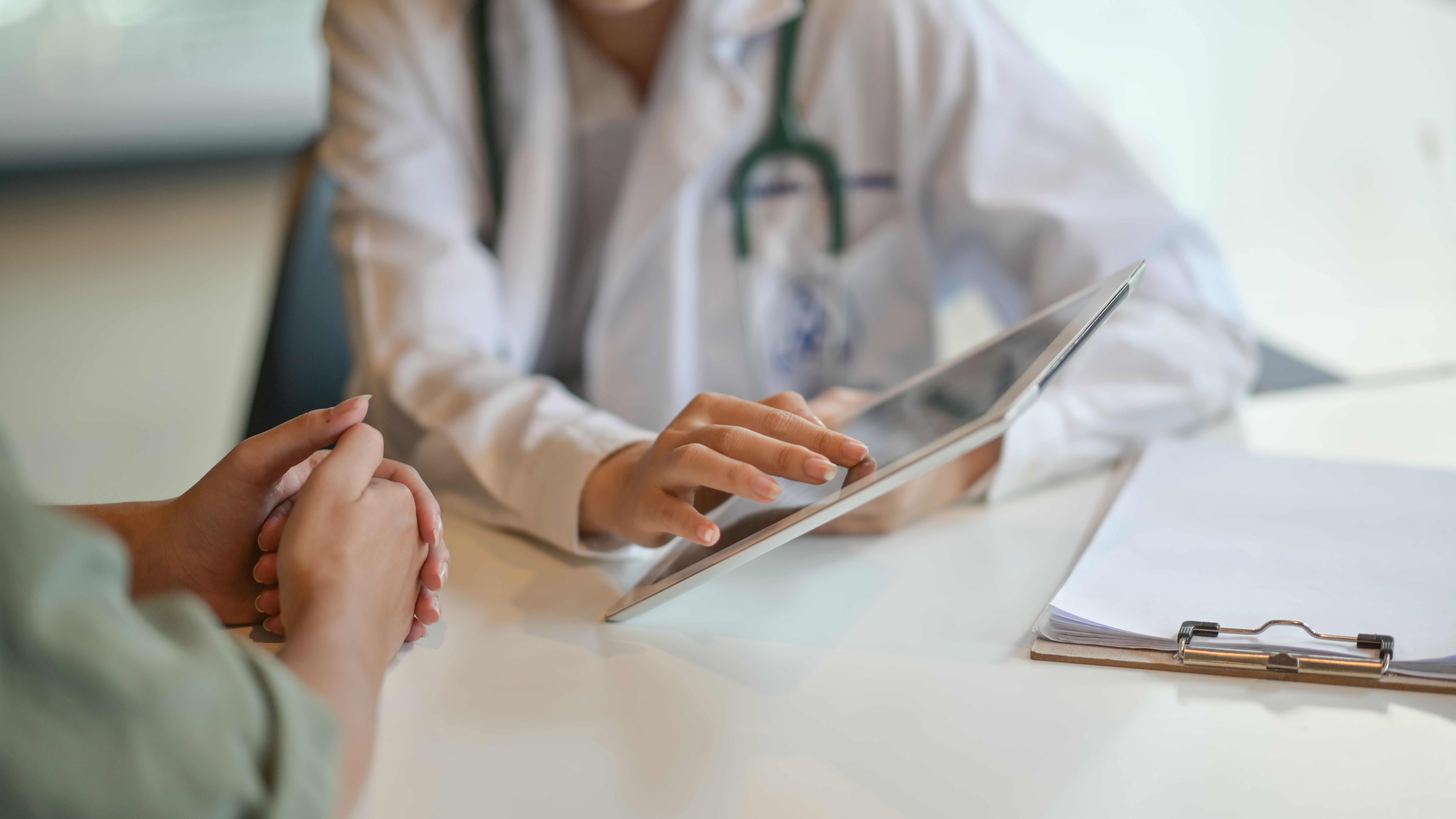 Close-up of a clinician using a tablet across a desk from an adult with clasped hands.